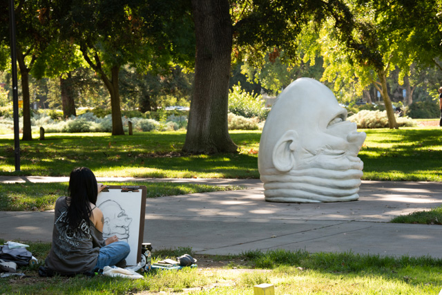 An artist, viewed from the back, looks on at the Eye on Mrak (Fatal Laff) Egghead as they sketch the sculpture in their notebook.