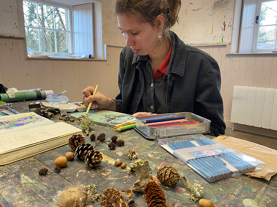 Class of 2023 alumna Ilsa Bauer draws from observation with pine combs, flowers and other pieces from nature in front of her on the table.l