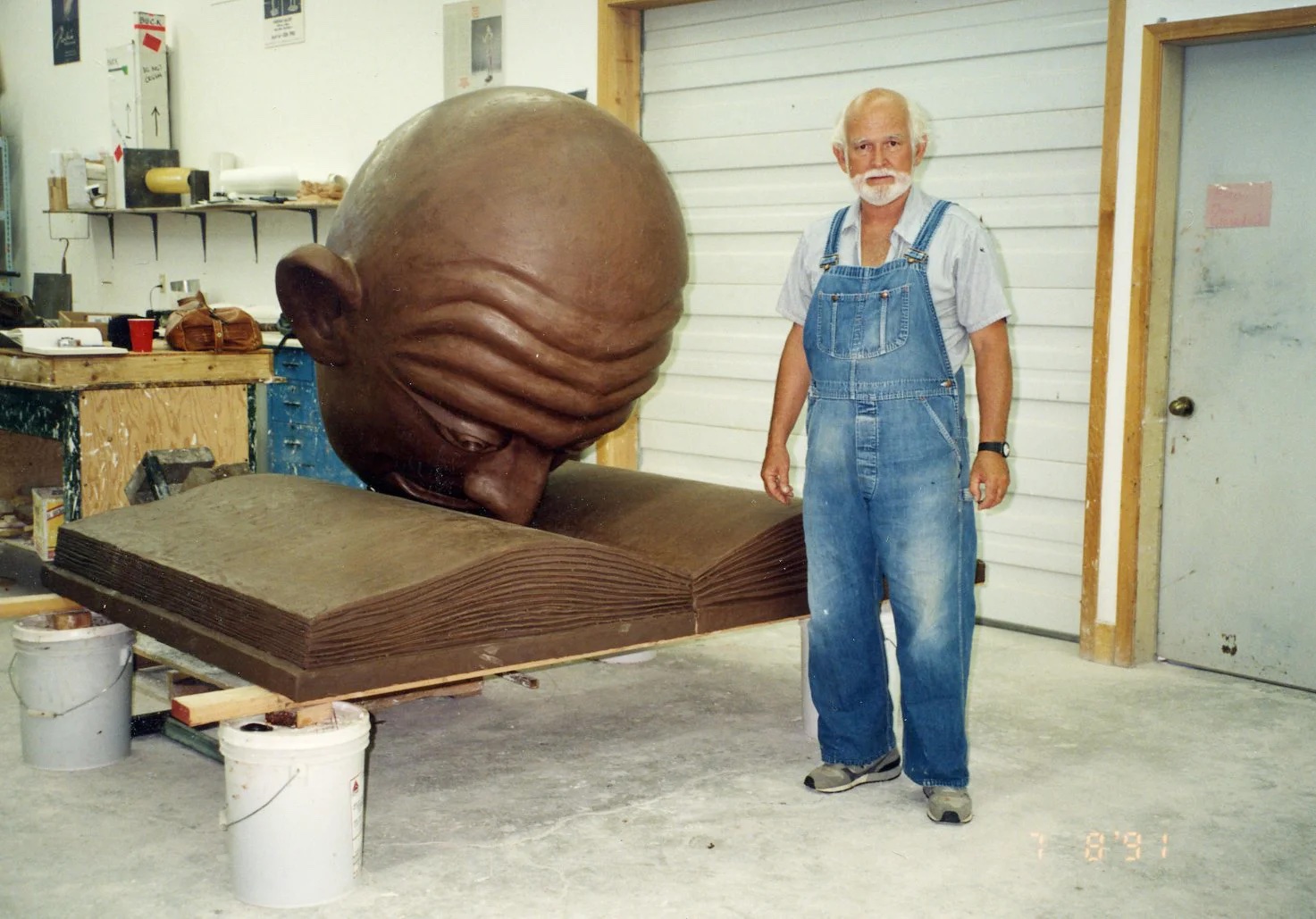 Robert Arneson, in denim overalls, with full-size clay mockup of Bookhead at Walla Walla Foundry. 