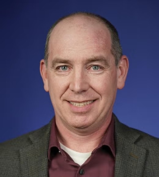 A man wearing a brown shirt and coat smiles at the camera