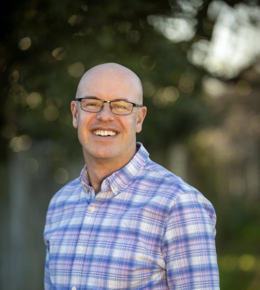 Smiling man in glasses wearing purple plaid shirt, outdoor photograph