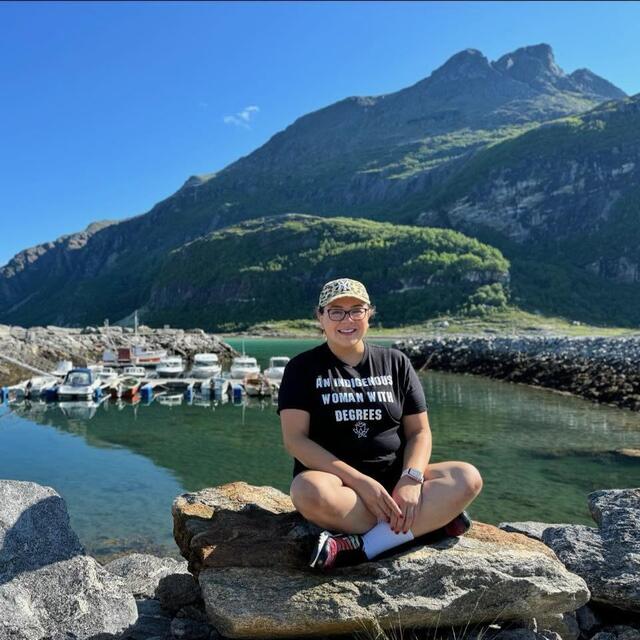 A woman sits cross-legged on a rock with a mountain and body of water behind her. Her t-shirt reads: "An Indigenous woman with degrees."