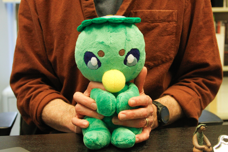 Professor Michael Dylan Foster holds a green stuffed kappa made by a student. The stuffy is between his hands, only his arms, hands and chest are visible behind the stuffed toy.