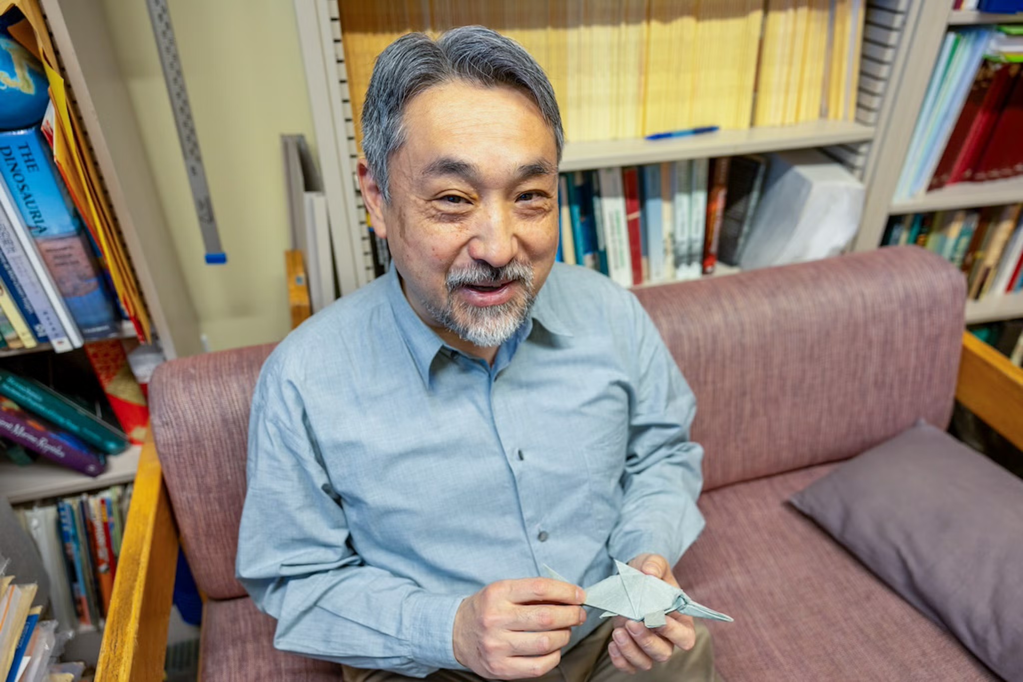 Older man in blue shirt smiling on couch, holding folded paper, bookshelves behind