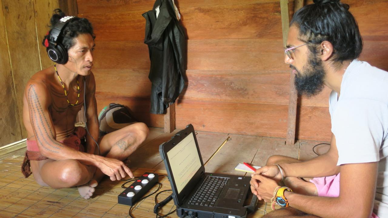 Two men sit on the ground, facing each other with one wearing headphones and the other looking at his laptop computer
