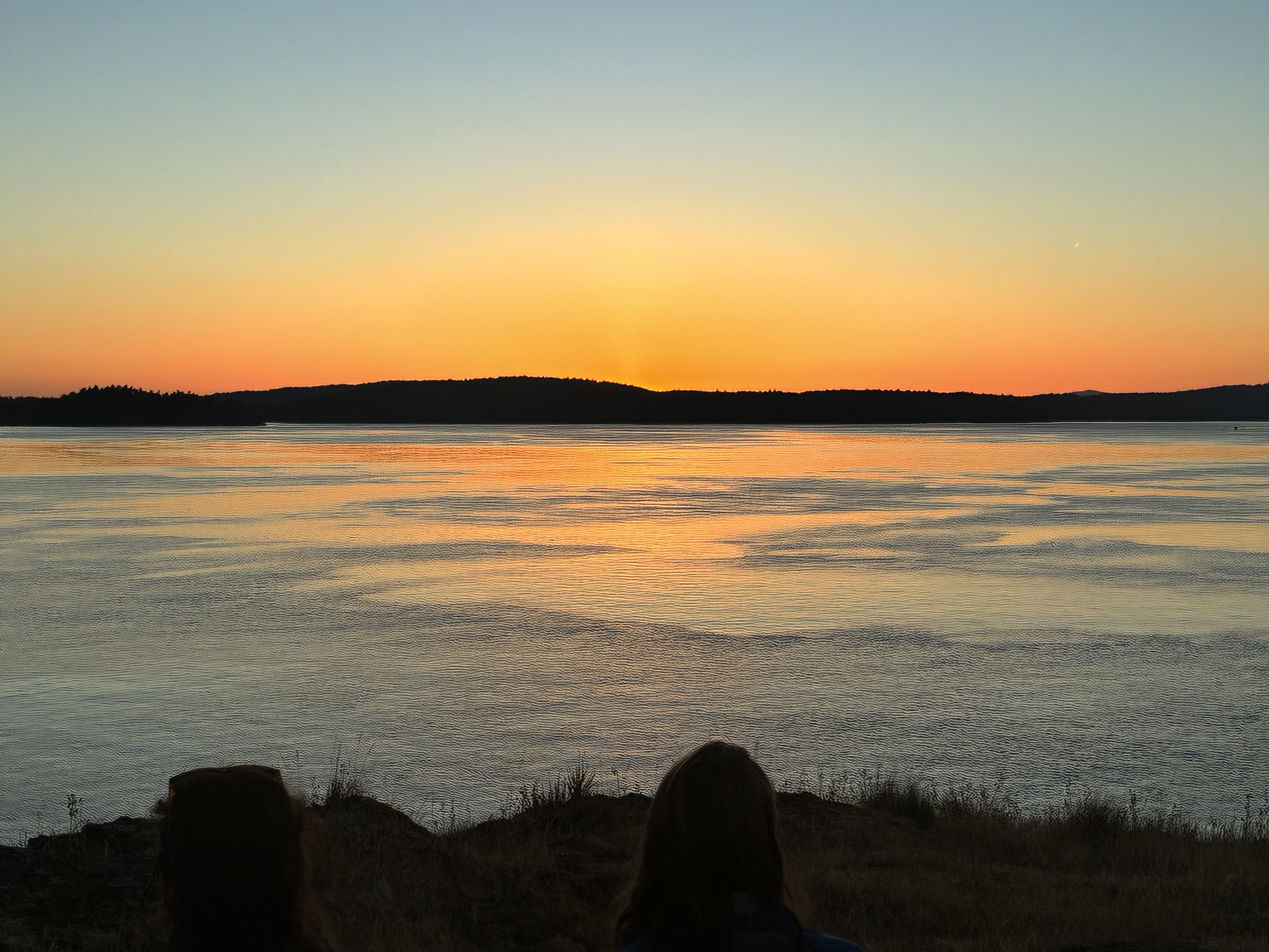Sunset over calm water with two silhouetted figures on a grassy shore