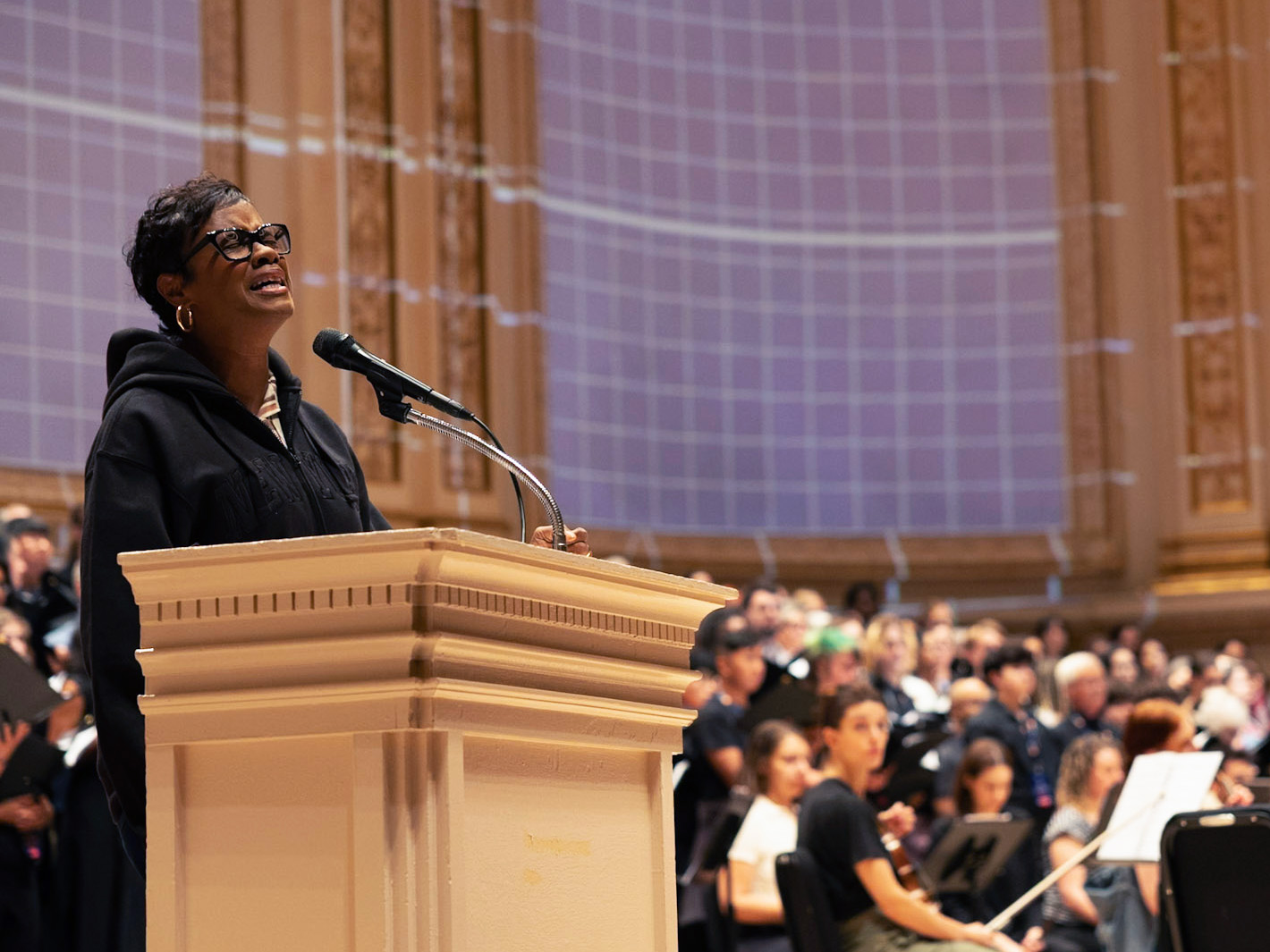 Woman speaking at podium, orchestra seated behind her in ornate concert hall