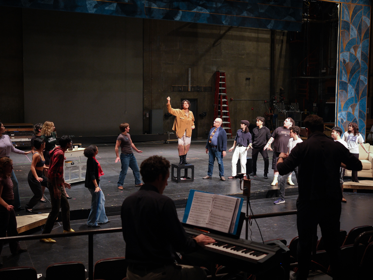 Actors rehearsing on stage with a conductor at a piano in the foreground.