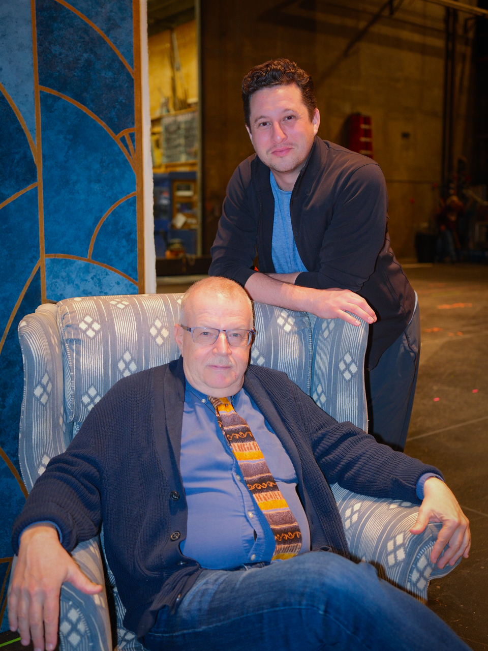 Two men pose together in a theater setting, one seated in an armchair and the other standing behind.