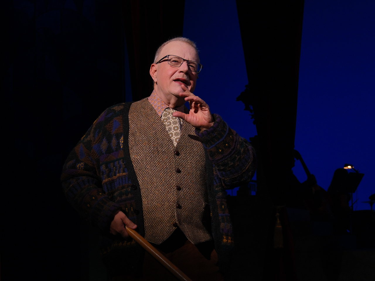 An older man in a sweater and vest gestures while speaking on stage with a blue backdrop.