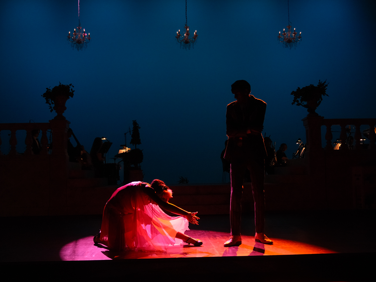 A dancer in a red dress reaches out to a man in a suit, illuminated by dramatic lighting.