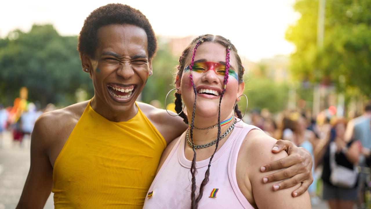 Friends hugging at outdoor festival; one in yellow, one in pink with rainbow face paint