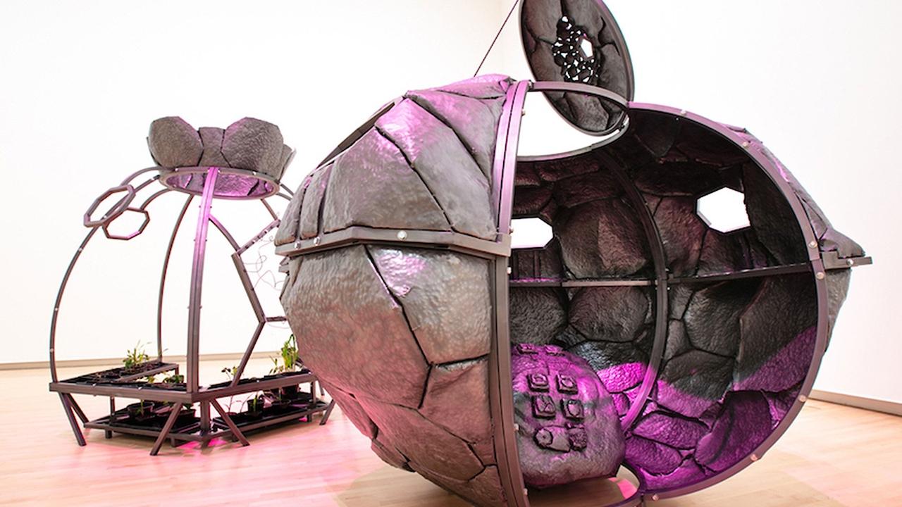 Industrial metal pod sculpture split open, purple-lit cushioned interior on gallery floor
