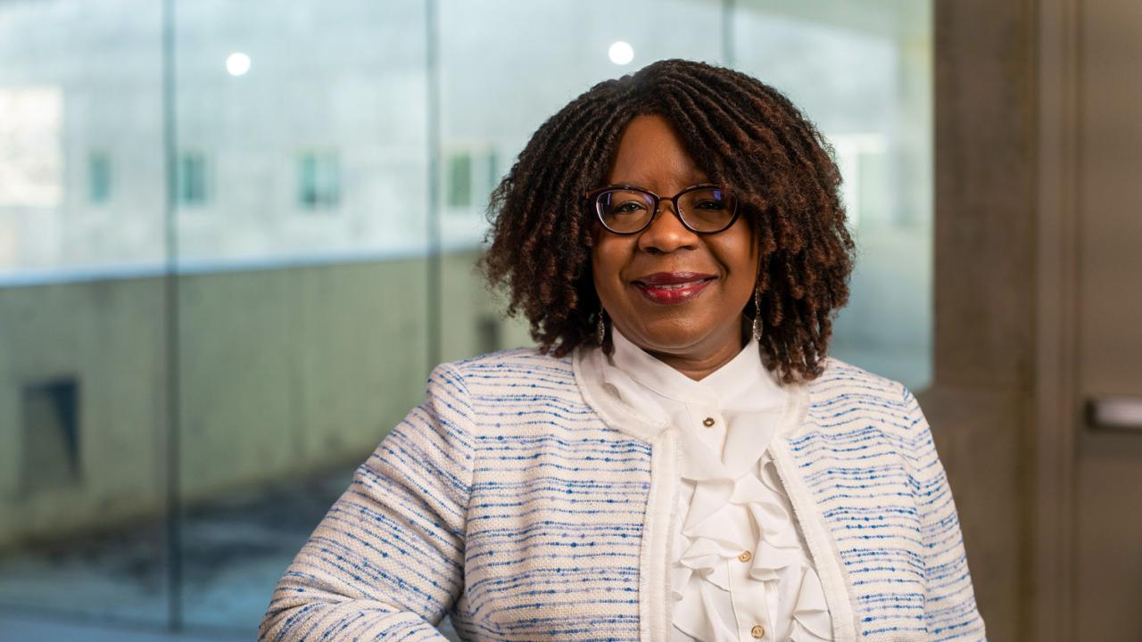 Portrait photo of a smiling woman seated in an office, wearing glasses and blue tweed jacket