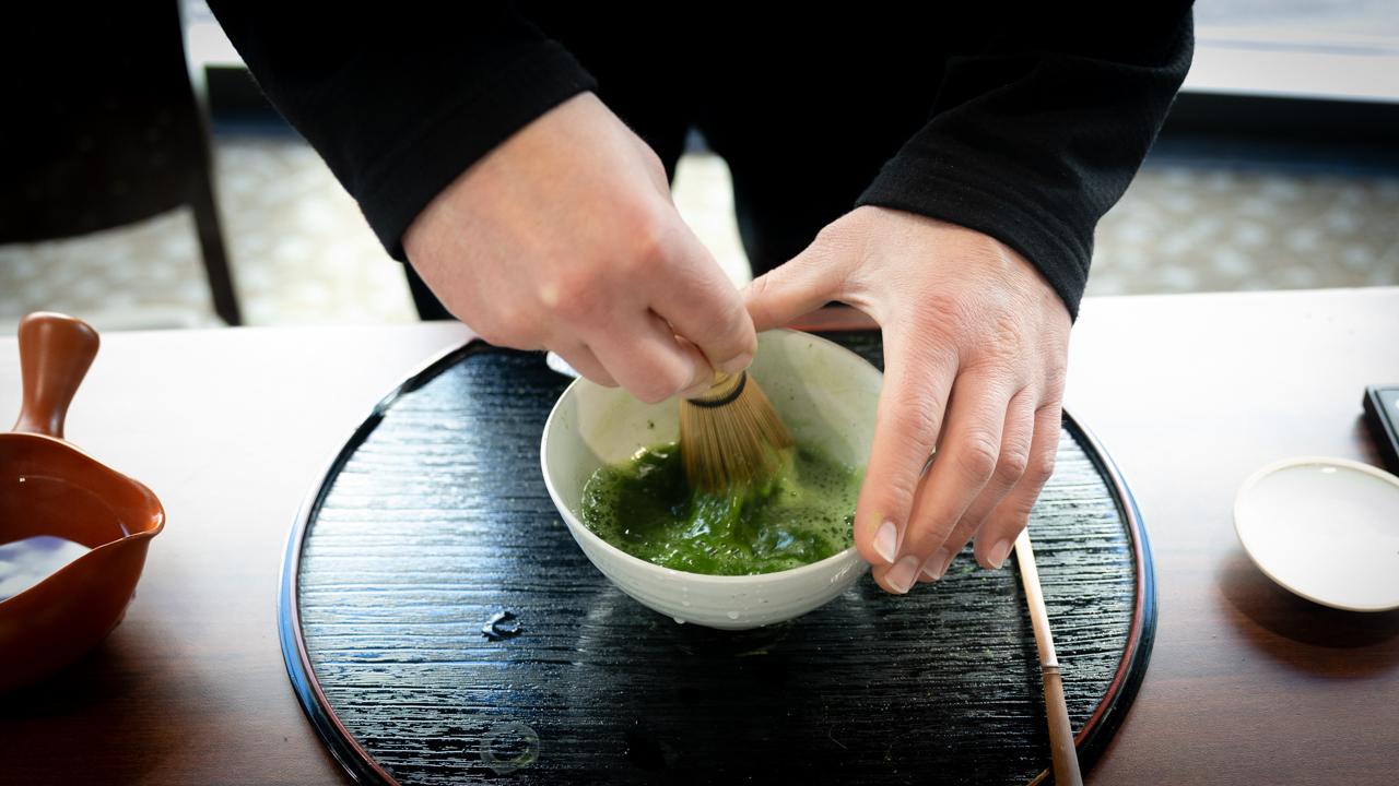 using a brush and bowl, two hands stir green matcha powder in liquid 