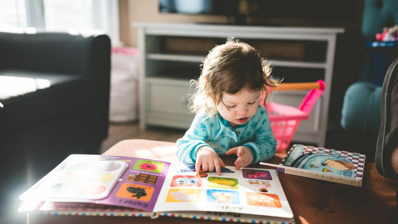 a toddler reading from a picture book on a couch.