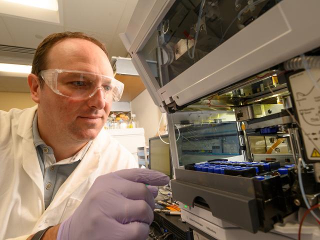 Scientist in lab coat, goggles and purple gloves adjusting analytical instrument