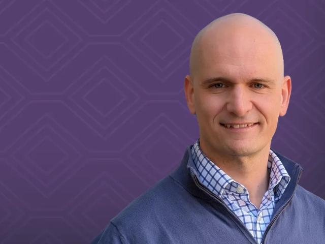 Smiling man wearing blue sweater and checked shirt, purple patterned background (photograph)