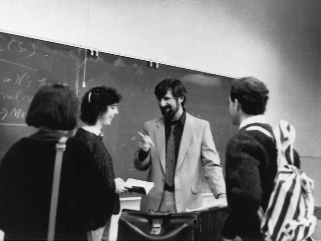 A professor engages with three students in a classroom, with a chalkboard in the background.