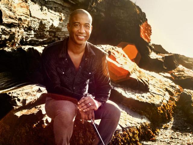 A man, Nicolás Dosman, sits on rocks smiling and looking into the camera with a sunburst effect over the image.