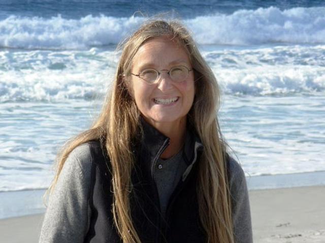 Smiling woman with long hair on a beach, waves and cloudy sky behind her.