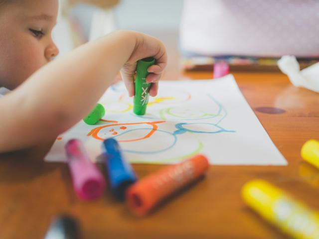 Focused toddler coloring with green crayon, colorful crayons scattered on wooden table