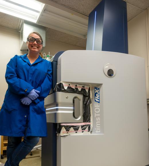 Elizabeth Neumann wears a blue coat, gloves and safety glasses as she stands next to Bruce, a matrix-assisted desorption/ionization mass spectrometer, or MALDI-MS.