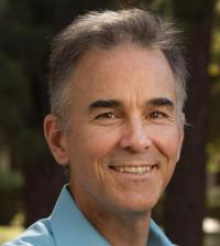 Smiling man with short gray hair, wearing a light blue shirt, in a natural outdoor setting.