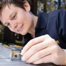 a person with short hair is wearing a turquoise green watch and handling a bee in a square tube. 