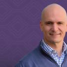 Smiling man wearing blue sweater and checked shirt, purple patterned background (photograph)