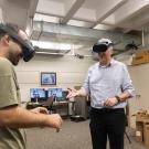 Two people wearing virtual reality headsets stand across from one another with their hands at waist level. Computers and boxes dot the background. 