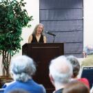 A woman speaks at a podium in front of an audience, with a potted plant nearby.