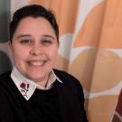 Smiling headshot of person with short dark hair, embroidered collar, abstract curtain background