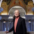 An older Chinese man posing for a photo in the great hall of the Library of Congress. He is wearing a dark pink dress shirt, black suit jacket and glasses.