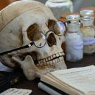 A skull with glasses resting on a wooden table near glass jars and an old pharmacy book.